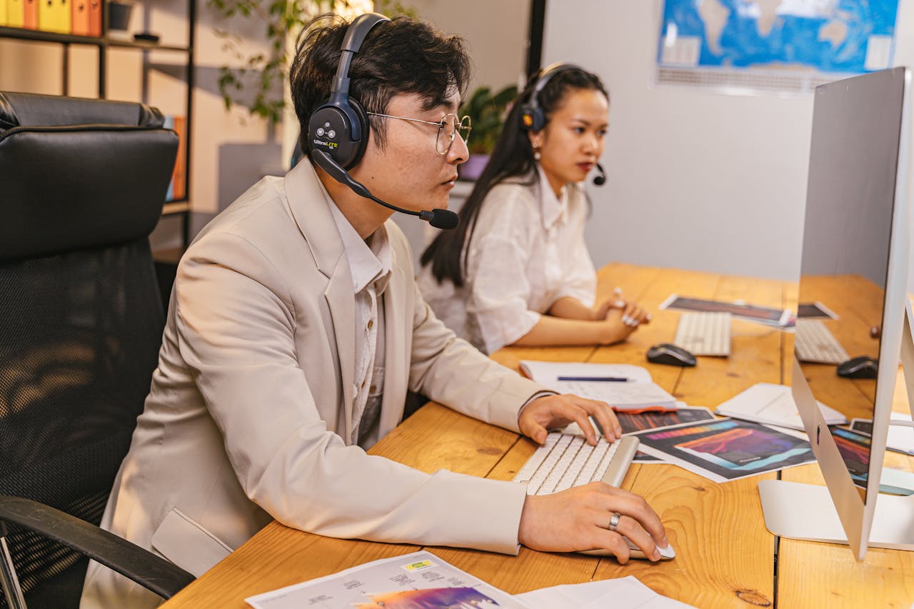 Two office workers using computers and headsets in a modern workspace, collaborating on tasks.